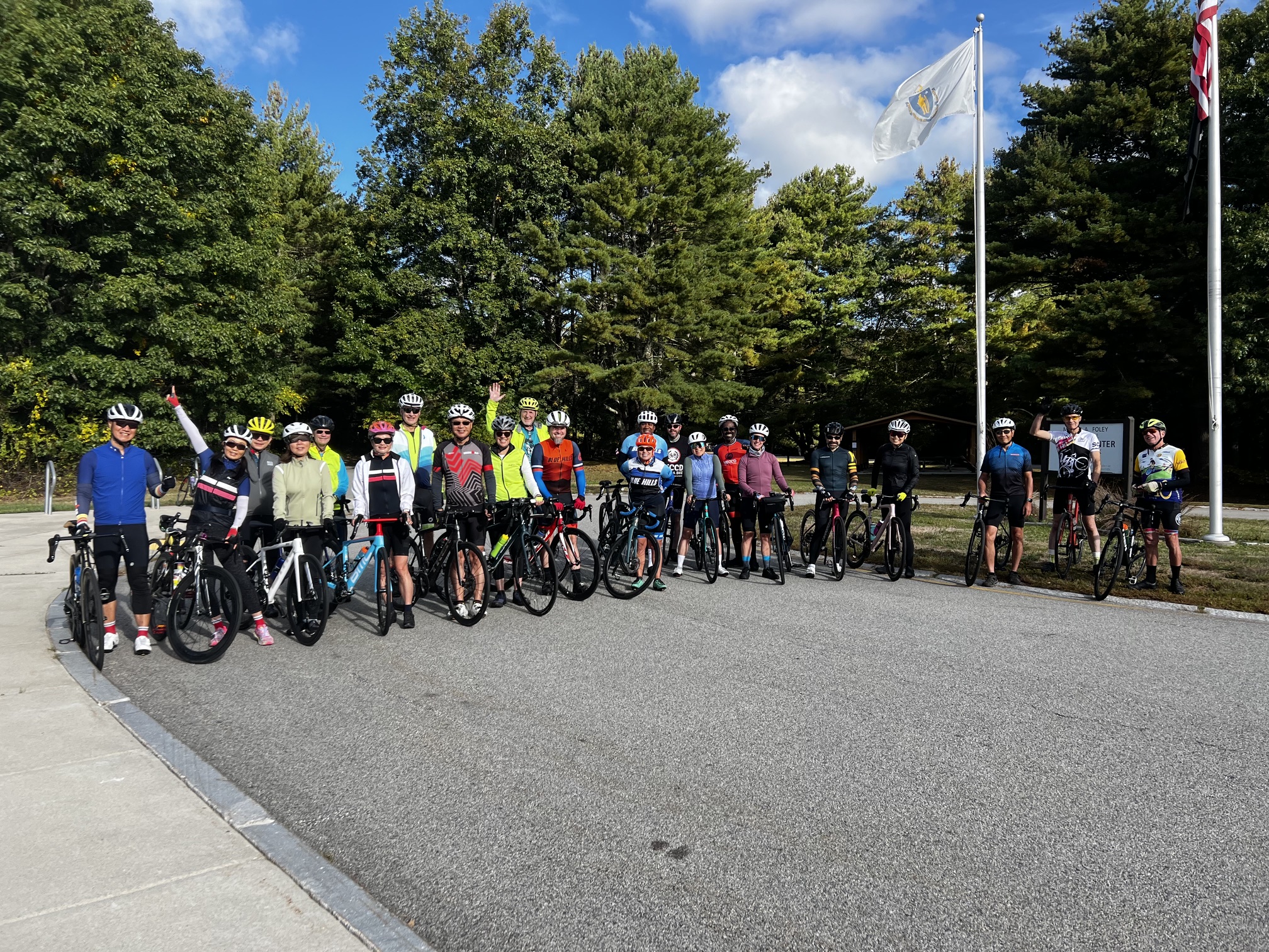 Cyclists stand with their bicycles in a broad group outdoors, with trees and flagpoles behind them; several wave toward the camera.